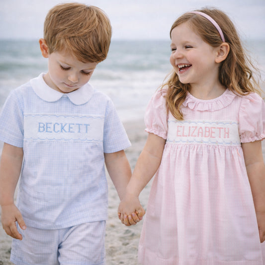 Two children, a boy and a girl, holding hands on a beach with smocked names on their clothing.