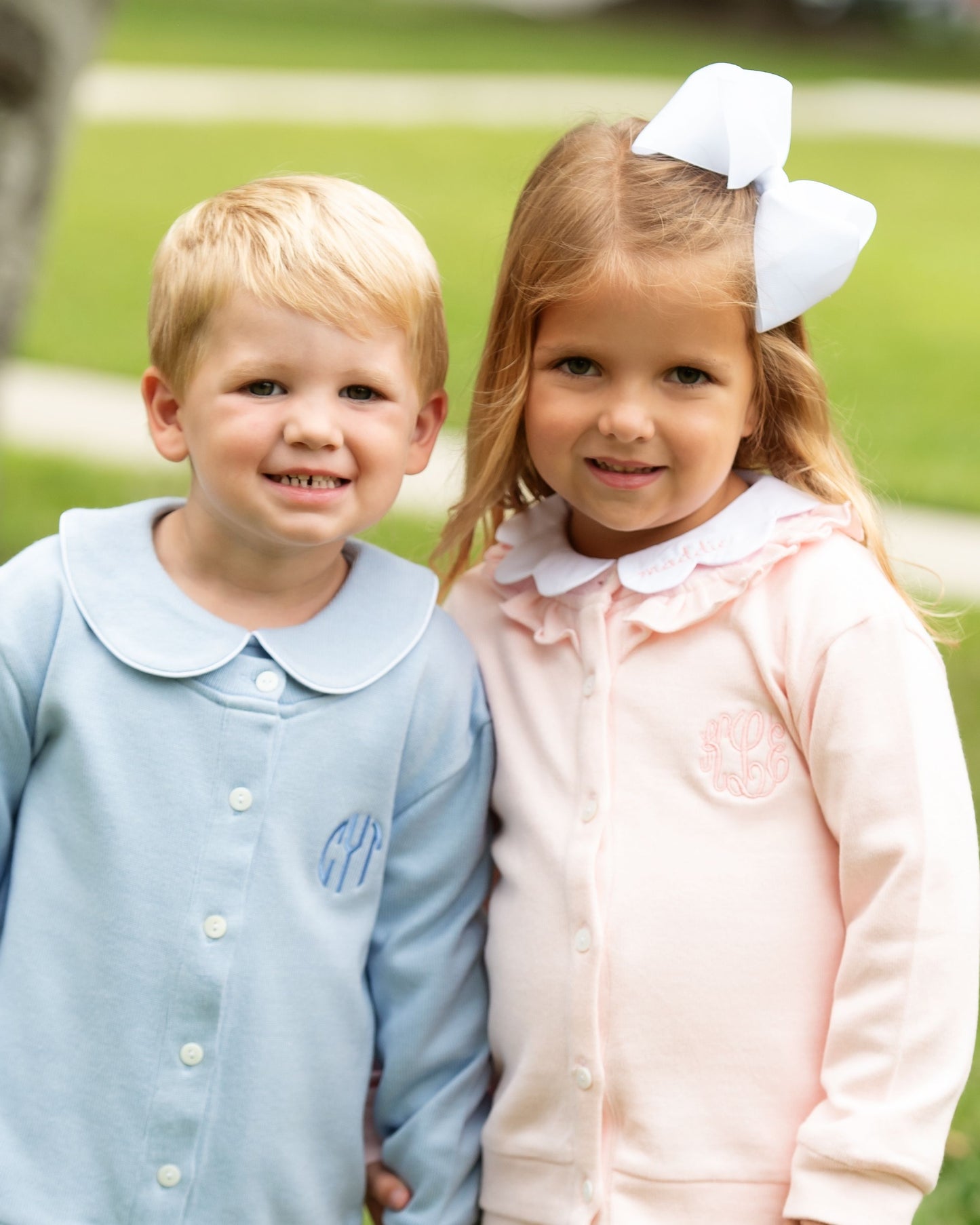 Two children, a boy and a girl, standing together outdoors with green grass in the background.