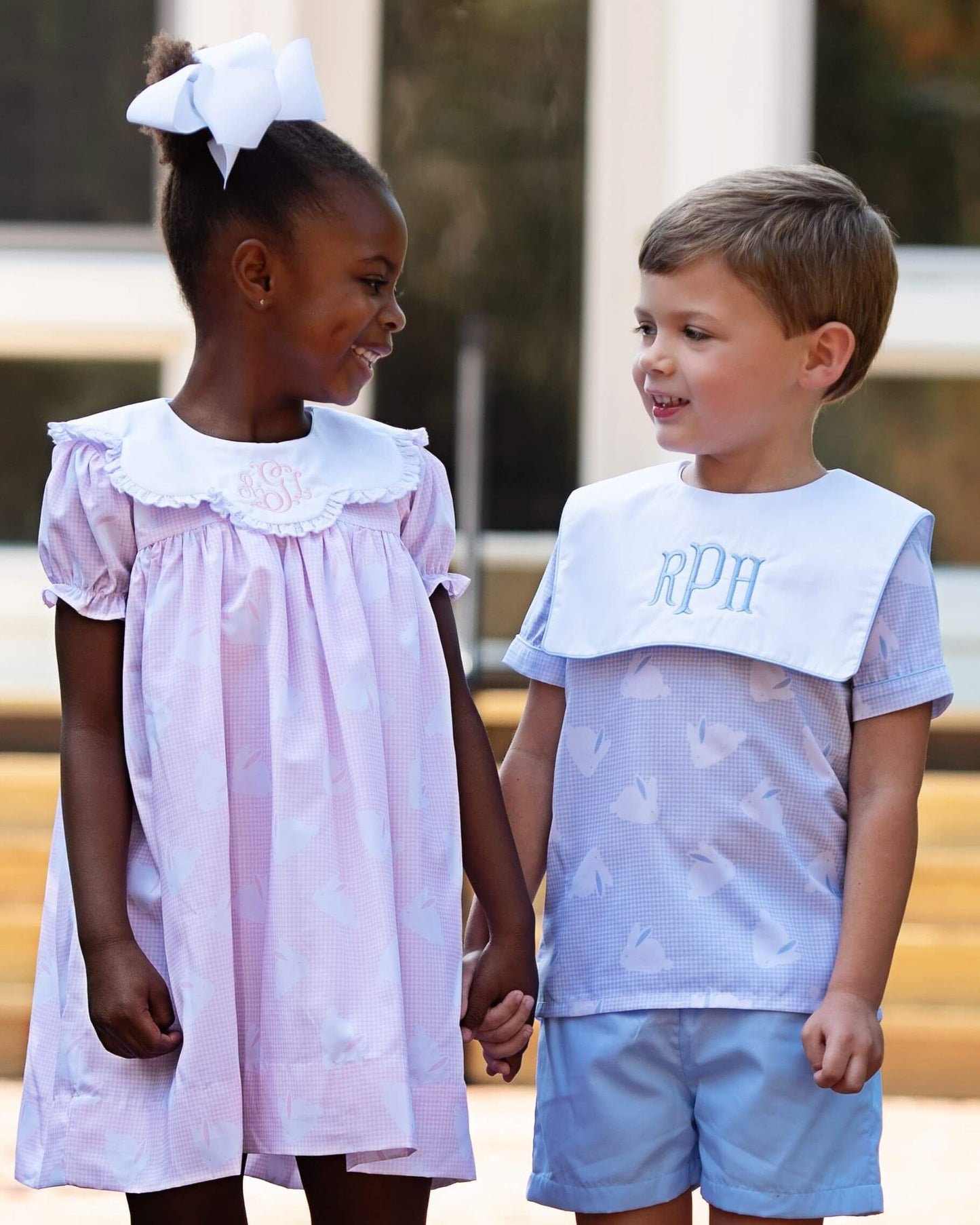 Two children, a girl in a light purple dress and a boy in a white shirt with 'RPH' and light blue shorts, standing outdoors.