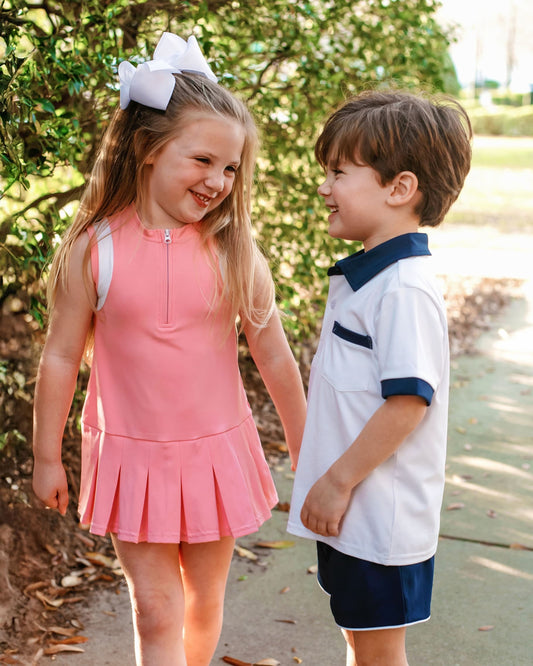 Two children, a girl in a pink dress and a boy in a white and navy outfit, standing outdoors with trees in the background.