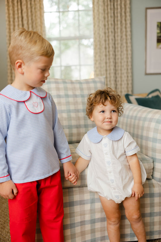 Two children holding hands in a living room with plaid curtains and a sofa.