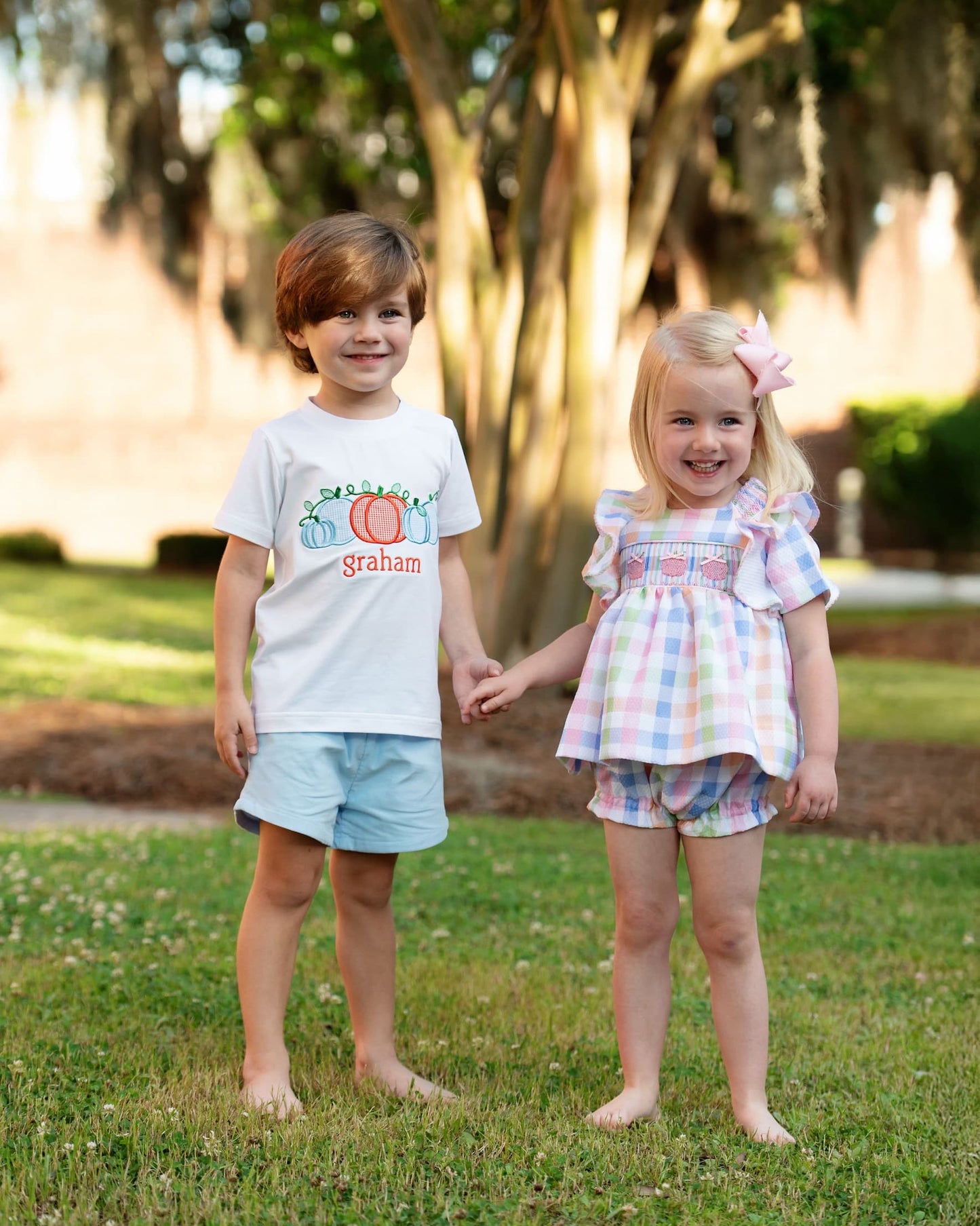 Two children holding hands in a park with trees and grass in the background.