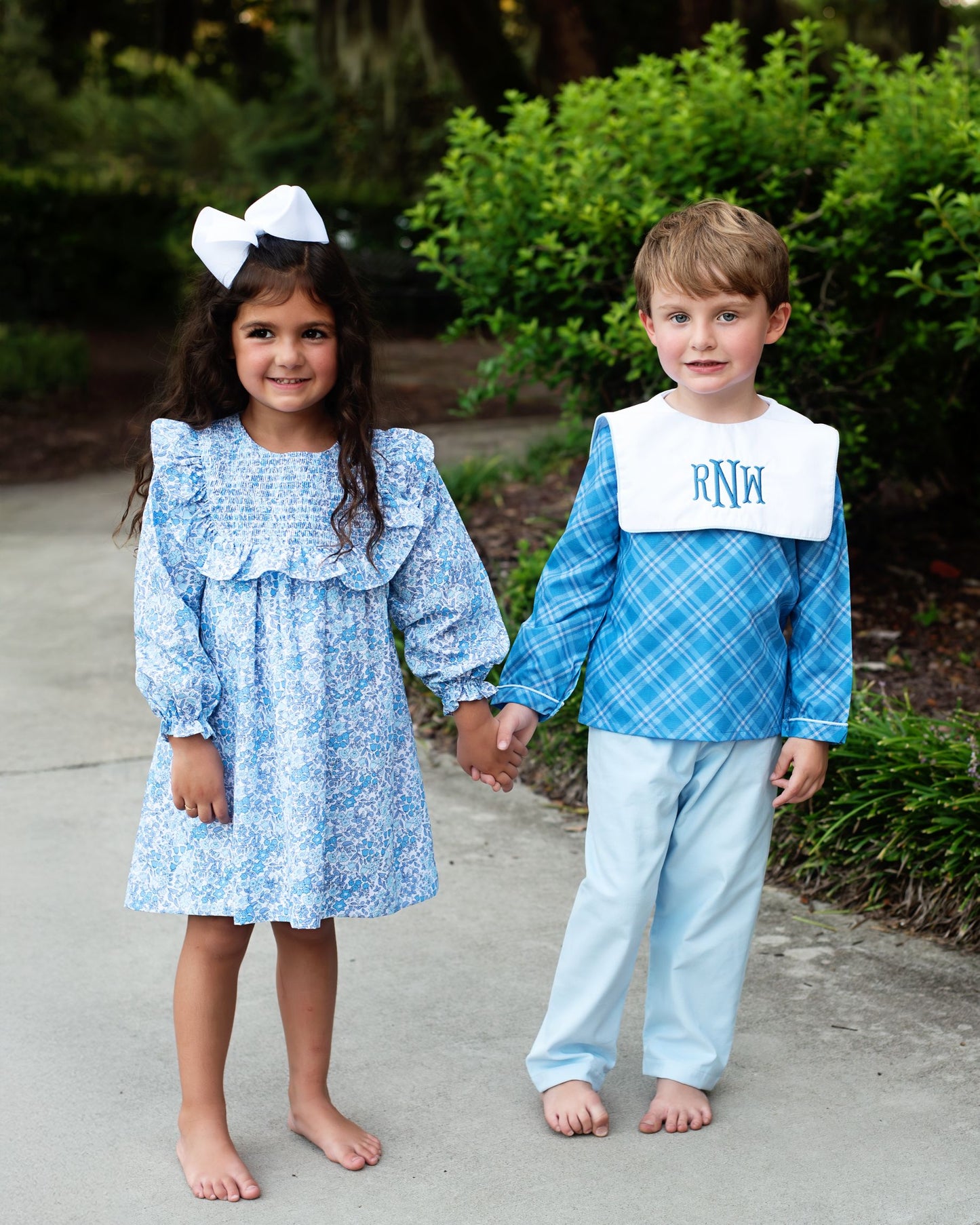 Two children holding hands, one in a blue dress with a white sailor hat and the other in a blue checkered shirt and pants with a bib, outdoors.