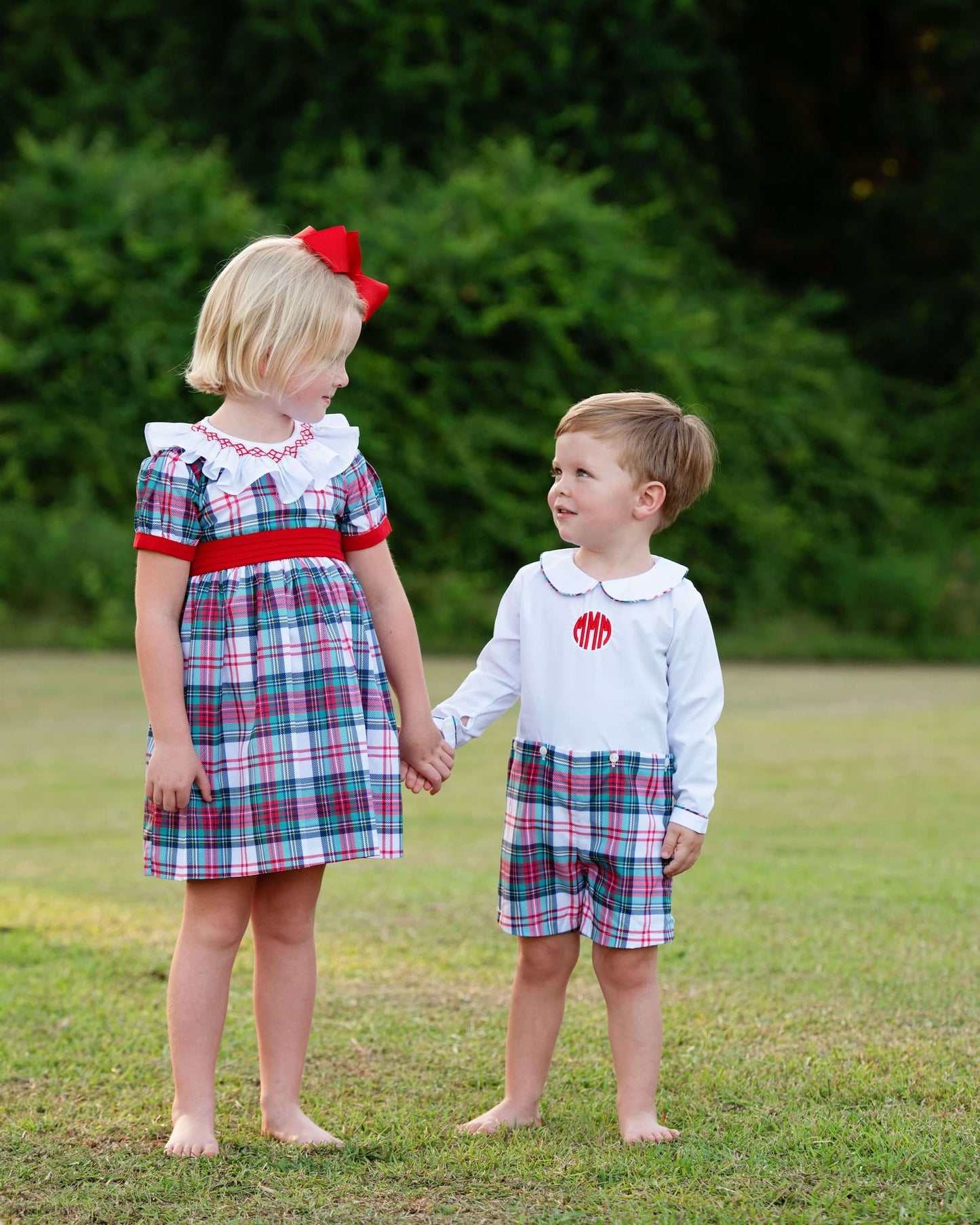 Two children holding hands outdoors, wearing matching plaid outfits with red accents.