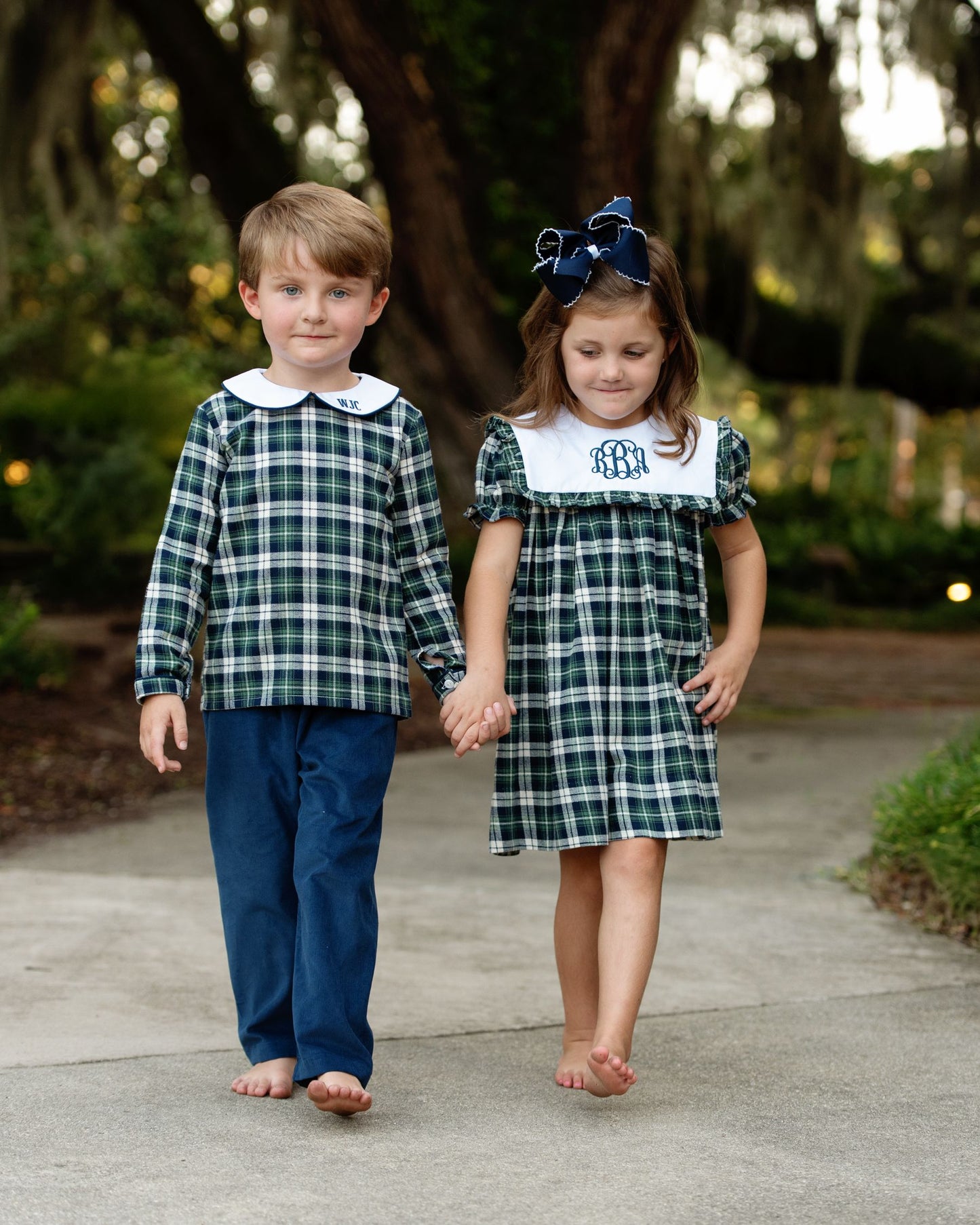 Two children in matching green plaid outfits walking hand-in-hand outdoors.