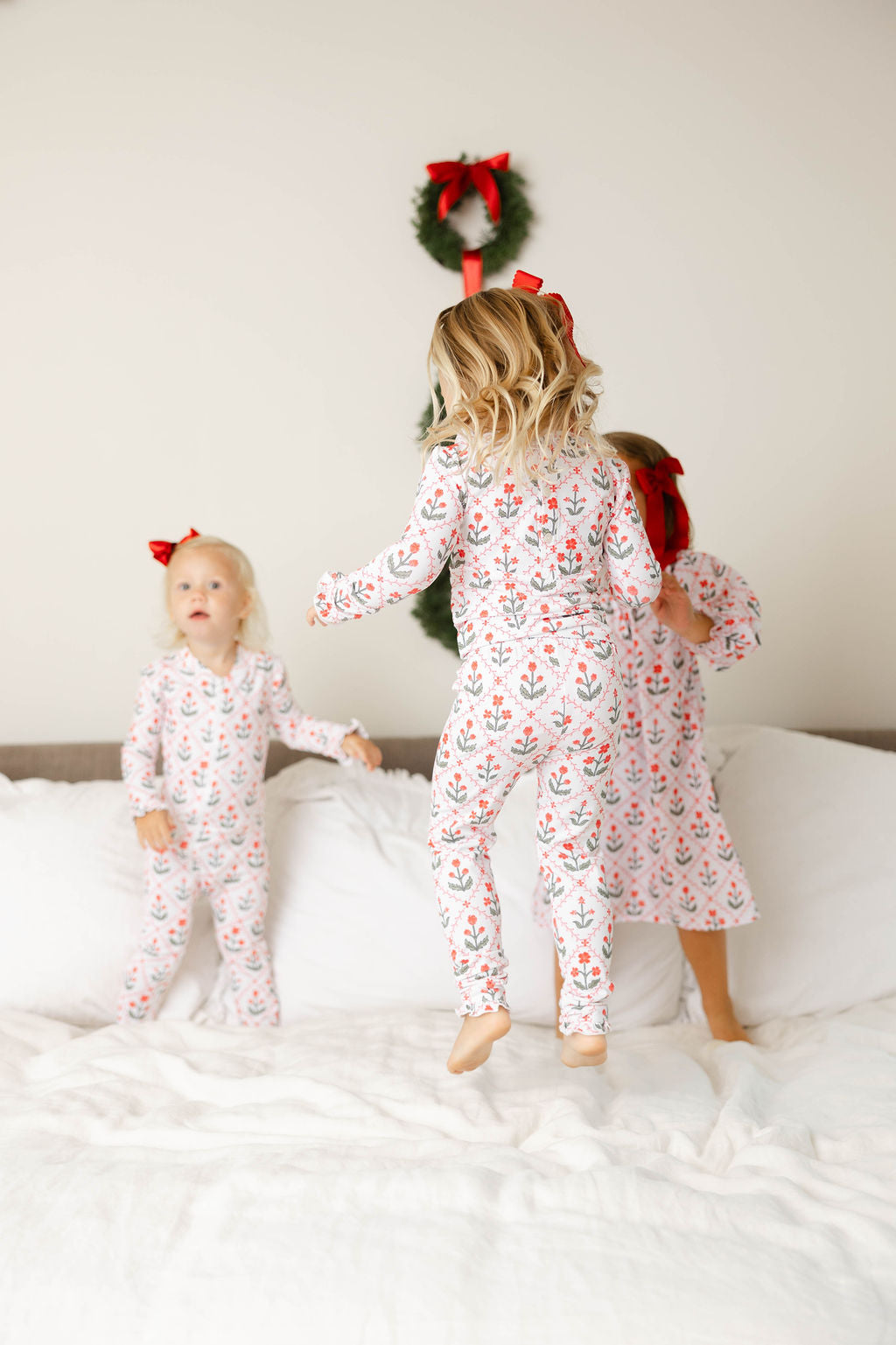 Two children in matching pajamas on a bed with a Christmas wreath in the background.