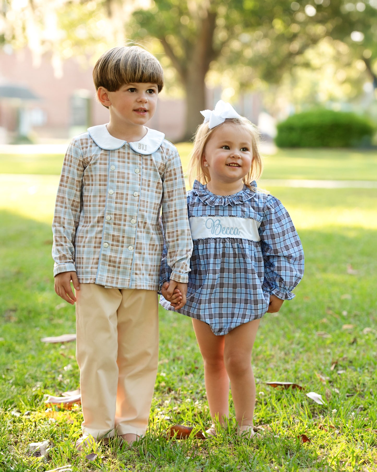 Two children in matching plaid outfits standing on grass with trees in the background