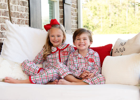 Two children in matching plaid pajamas sitting on a couch with pillows.
