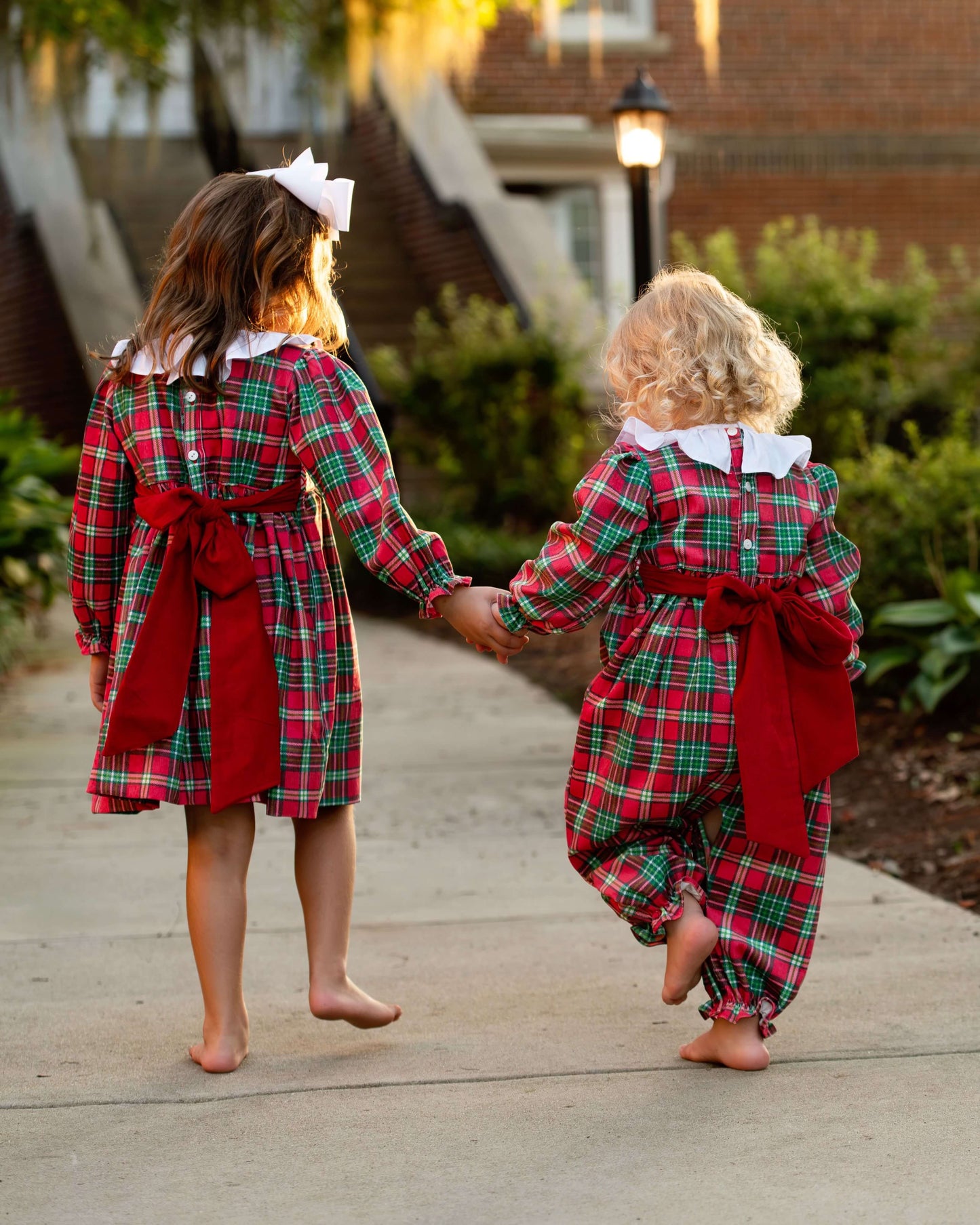 Two children in matching red and green plaid outfits walking hand in hand on a wooden deck.