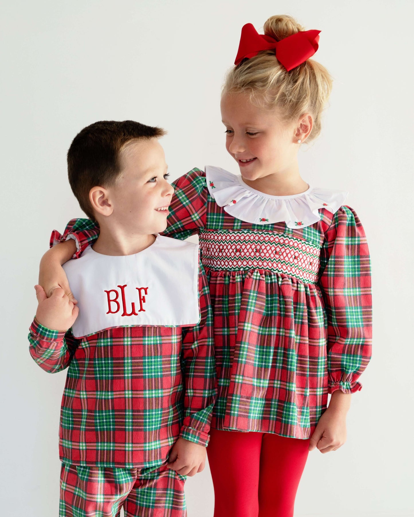 Two children in matching red and green plaid outfits with a white bib and red bow.