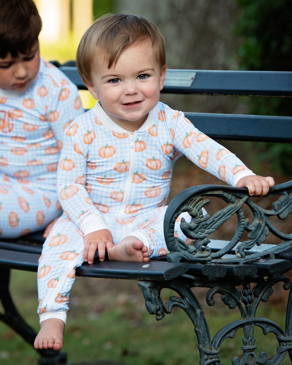 Two children in pumpkin-patterned outfits sitting on a bench outdoors.