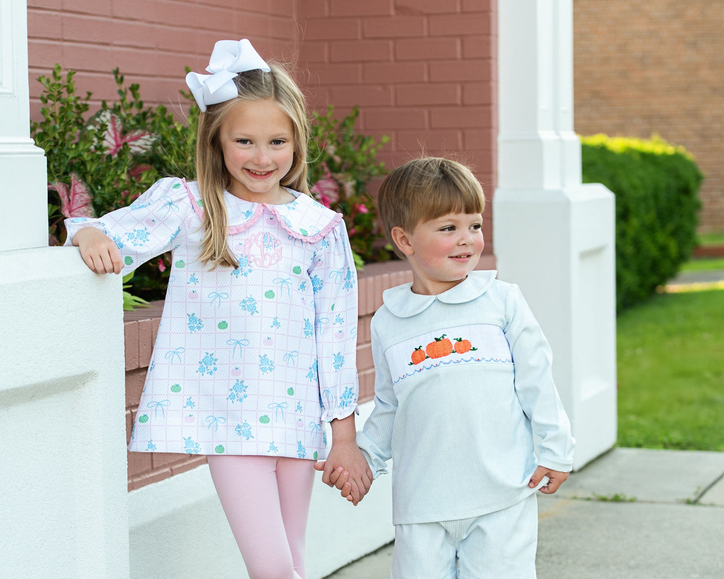 Two children in pumpkin outfits standing on a porch with a brick wall and greenery in the background 1