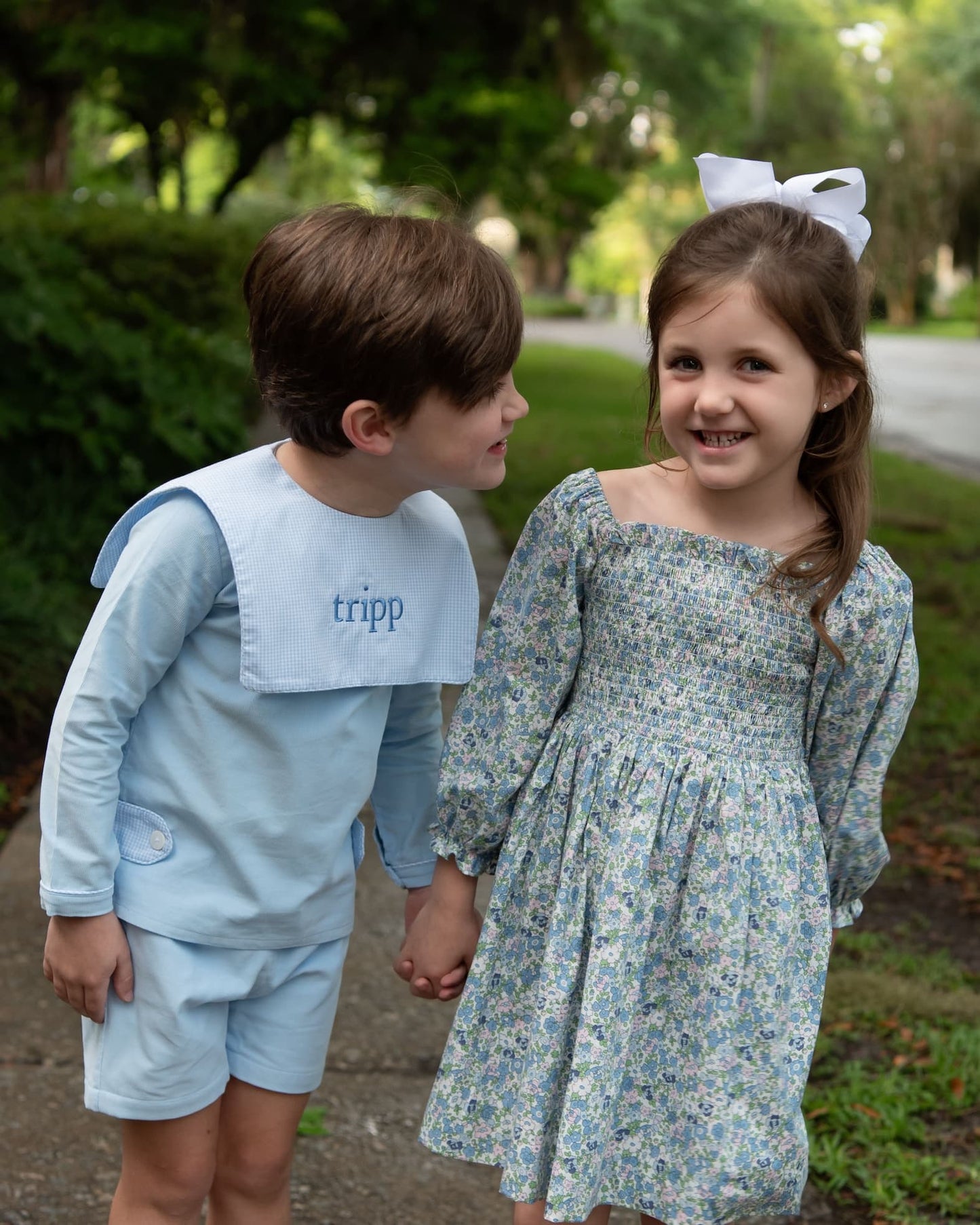 Two children outdoors holding hands, wearing matching light blue and floral printed outfits from The Tripp. The boy wears a light blue long-sleeve top and shorts with a white bib, and the girl wears a light blue floral long-sleeve dress with a white bow in her hair.