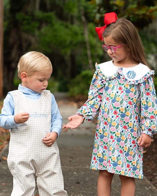 Two children outdoors on a path. The child on the left wears a light blue long-sleeve shirt and white gingham overalls with the name 'Molten' embroidered on the chest.
