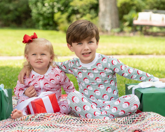 Two children sitting on a blanket outdoors, wearing matching holiday pajamas. The boy wears light gray pajamas with a red Santa Claus pattern, and the girl wears pink pajamas with red stripes and a red bow in her hair.