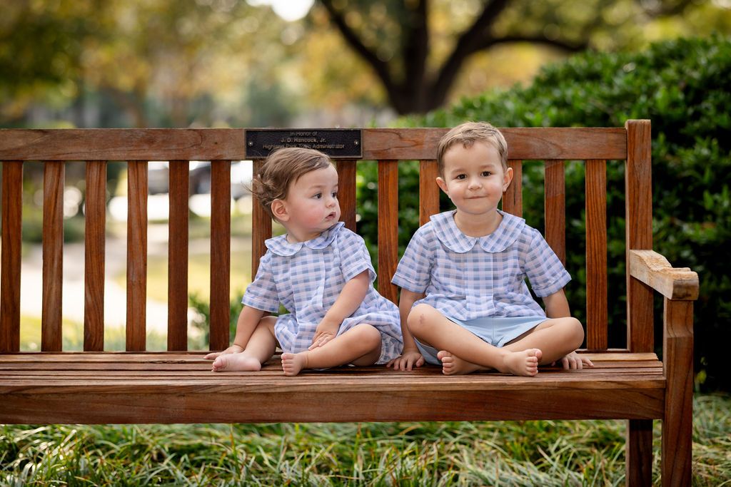 Two children sitting on a wooden bench outdoors with greenery in the background