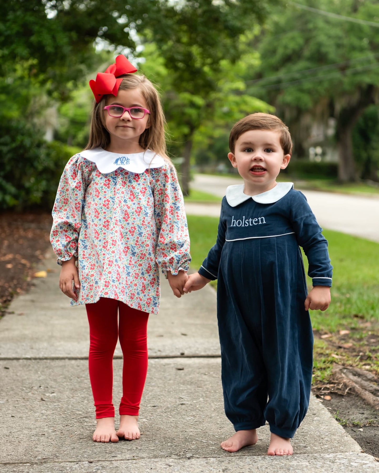 Two children standing on a sidewalk outdoors, holding hands. The girl wears a floral long-sleeve tunic with a white Peter Pan collar, red leggings, and a large red bow in her hair.