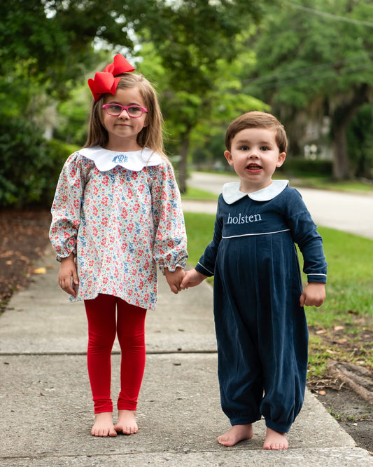 Two children standing on a sidewalk outdoors, holding hands. The girl wears a floral long-sleeve tunic with a white Peter Pan collar, red leggings, and a large red bow in her hair.