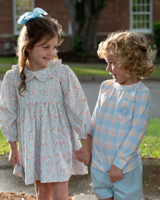 Two children standing outdoors and holding hands. The girl wears a light blue floral long-sleeve dress with a white collar and a blue bow in her hair. The boy wears a plaid shirt and blue shorts