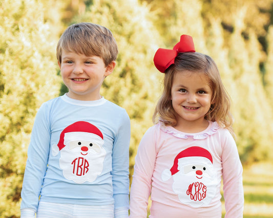 Two children standing outdoors in front of a blurred yellow-green background, wearing matching long-sleeve shirts with a Santa Claus graphic. The child on the left wears a light blue shirt with a white Santa graphic and red monogram '1BR', and the child on the right wears a light pink shirt with a white Santa graphic and red monogram 'MRO'.