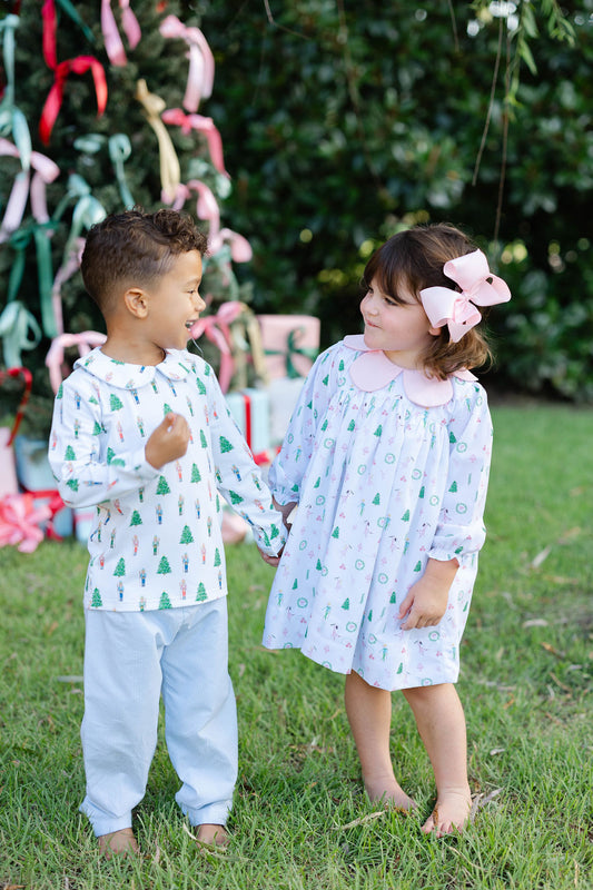 Two children standing outdoors, one in a patterned shirt and pants, the other in a dress with a bow, with a decorated Christmas tree in the background.