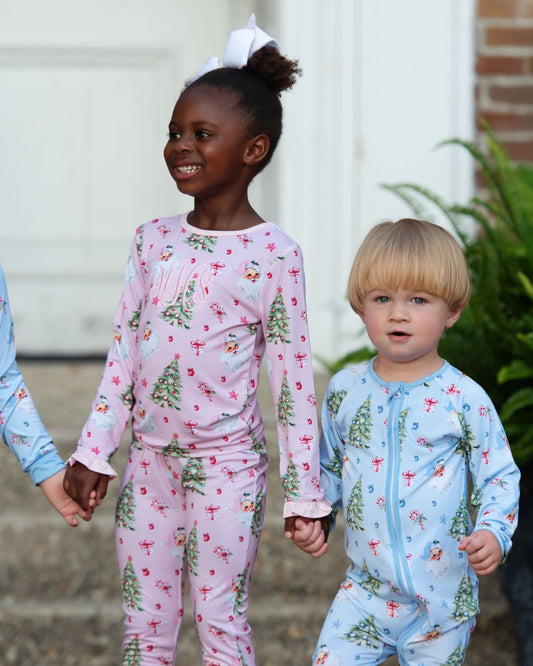 Two children wearing matching holiday-themed pajamas, with the girl in a pink long-sleeve pajama set and the boy in a light blue one-piece, both printed with Christmas trees and ornaments, standing outdoors and holding hands.