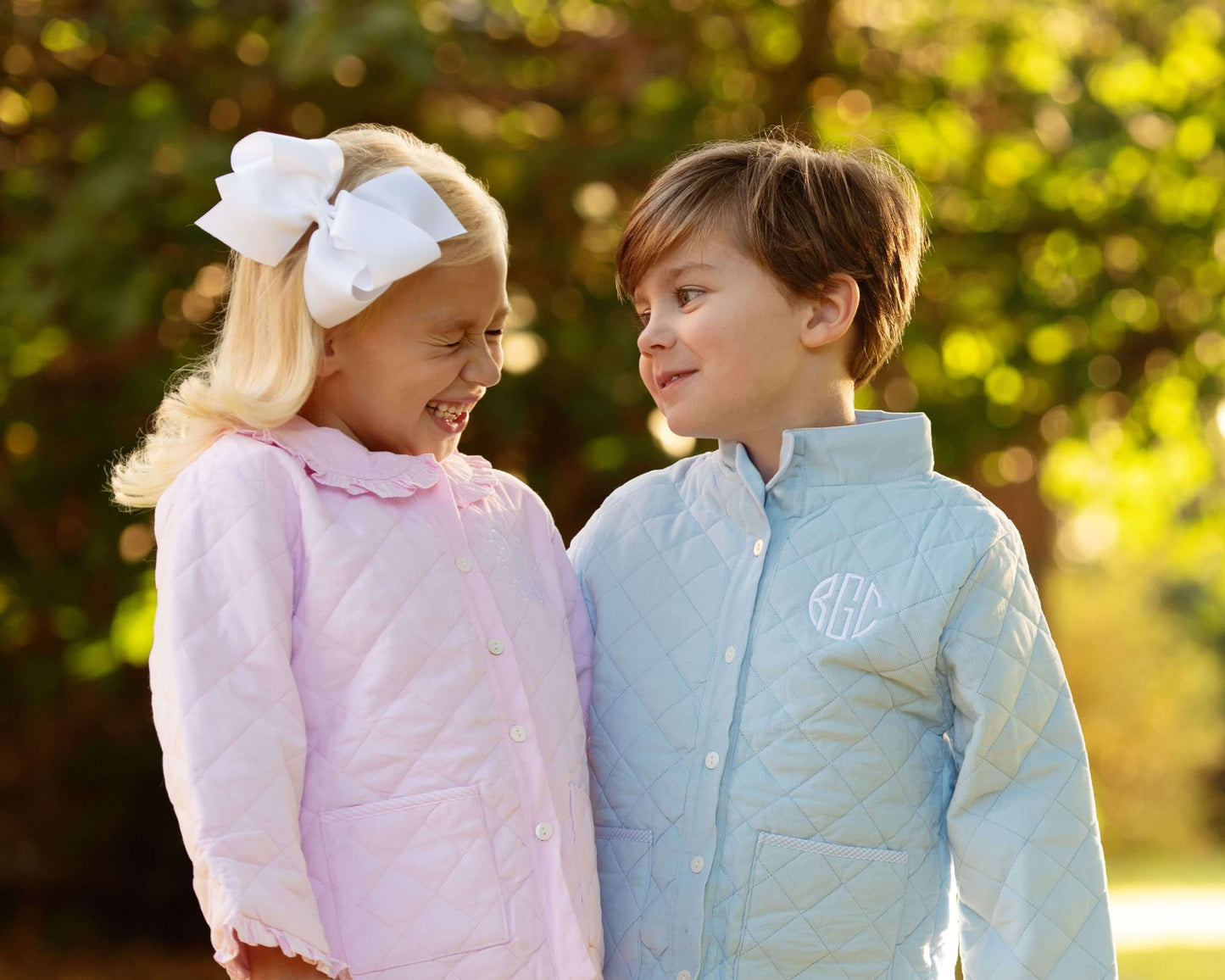 Two children wearing quilted jackets outdoors with a blurred natural background