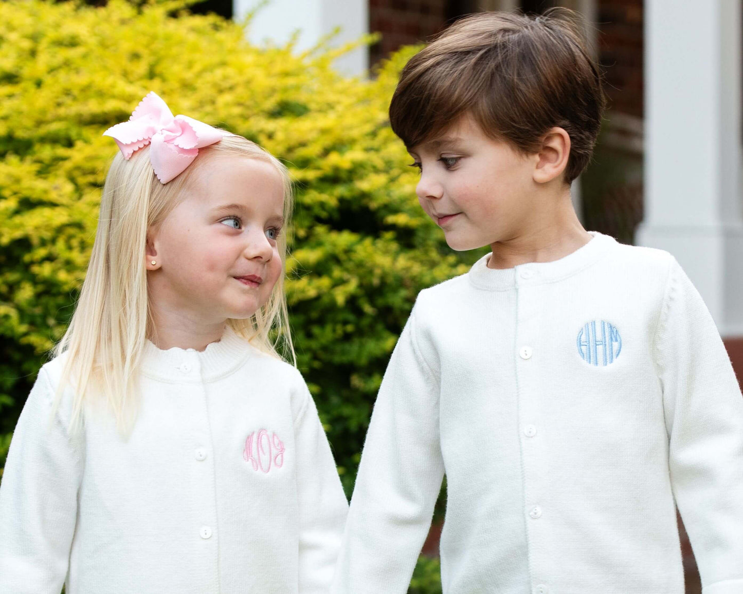 Two children wearing white jackets with monograms, standing outdoors.