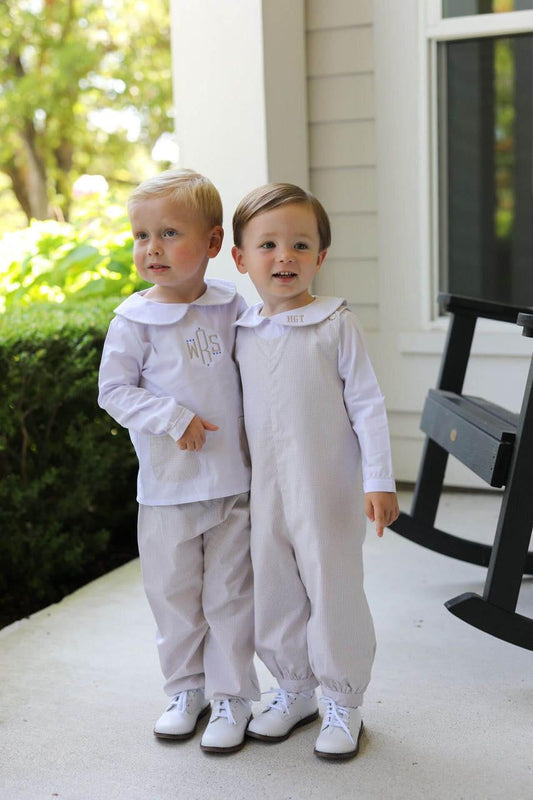 Two toddlers standing on a porch wearing matching white formal outfits, one long-sleeve top with matching pants and the other white jumpsuit, both with white shoes.