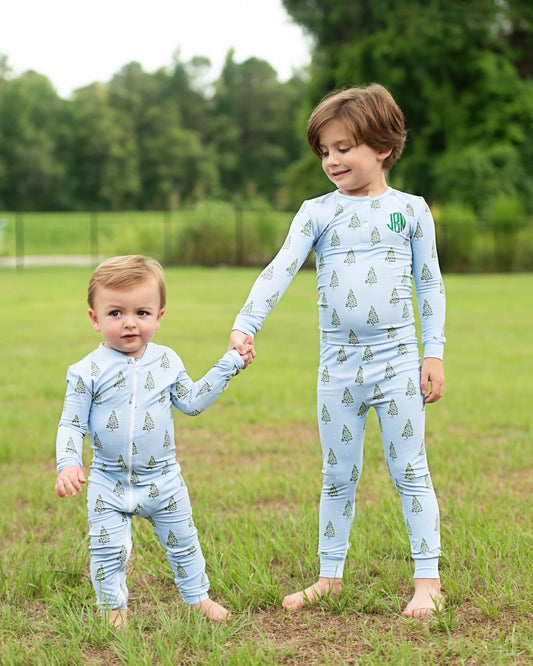 Two toddlers wearing matching light blue long-sleeve pajamas with a green pine tree pattern, standing barefoot in a grassy park. The older child has a green monogram on the chest.