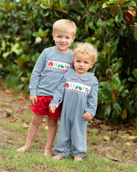 Two young boys standing outdoors on grass in front of dense green foliage, wearing matching long-sleeve striped pajamas with a smocked tractor design on the chest. The older boy also wears red shorts.