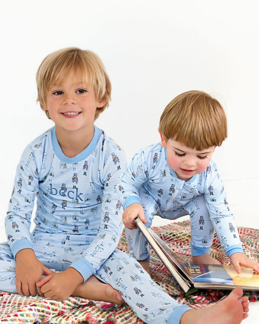 Two young boys wearing matching light blue long-sleeve pajamas with a small bear print and the word 'back' on the chest, sitting on a patterned blanket and reading a book.