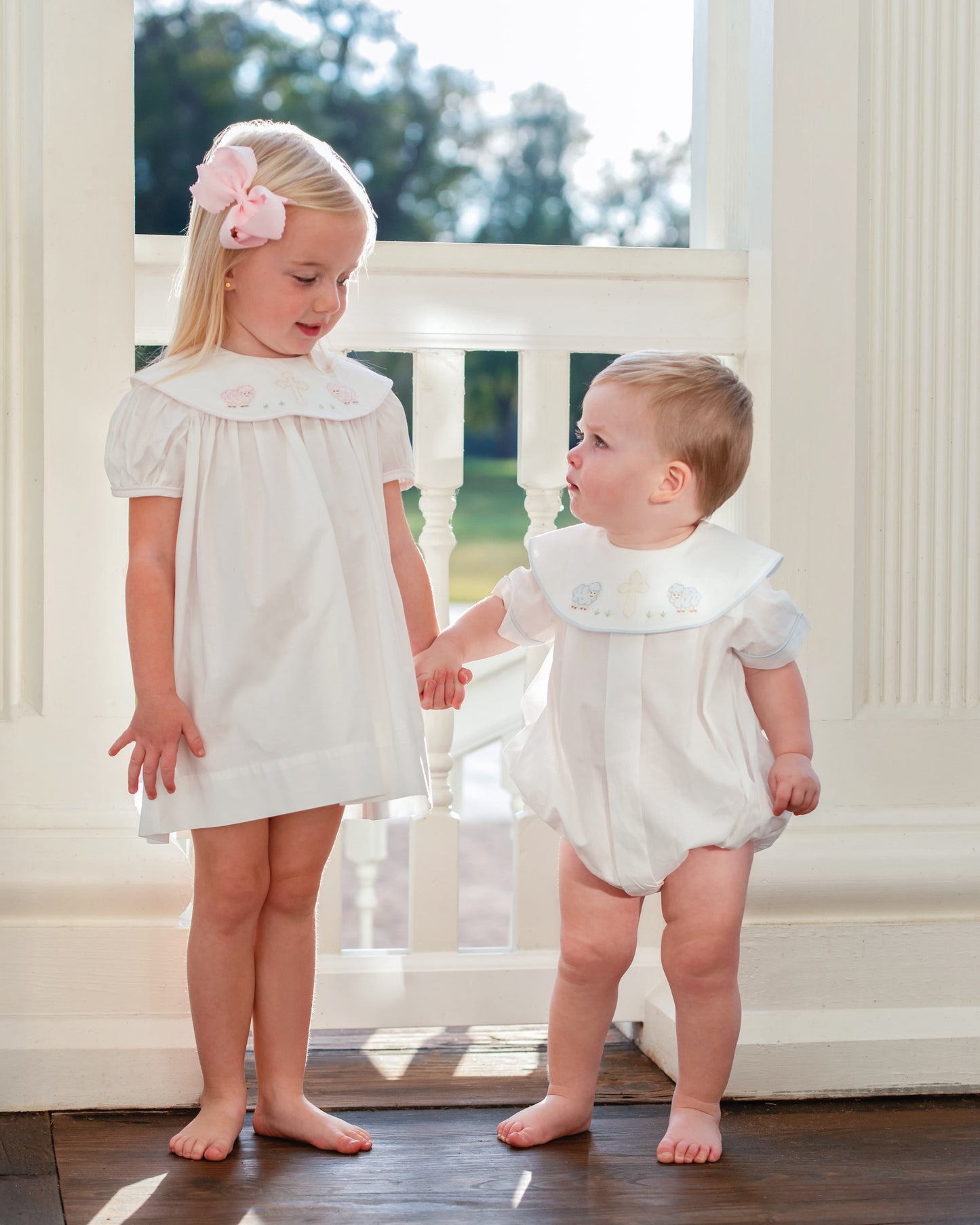 Two young children in white outfits standing on a wooden porch.