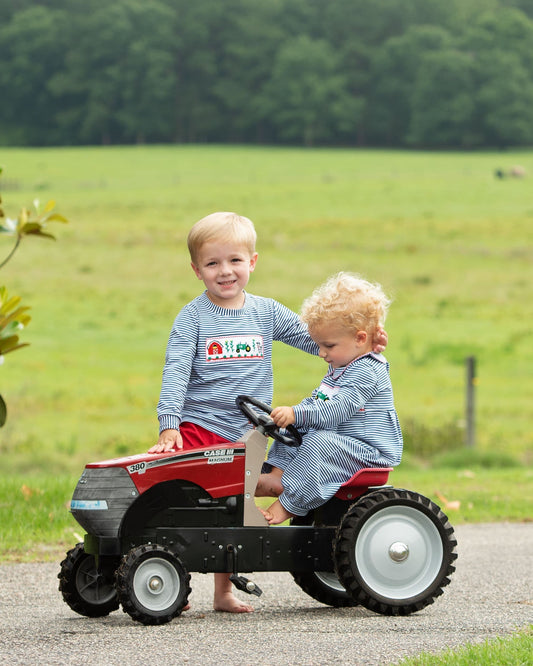 Two young children outdoors on a paved path in a grassy field, with one sitting on a red and black toy tractor and the other standing beside it. Both are wearing matching blue-and-white striped long-sleeve shirts and light blue striped pants or shorts.