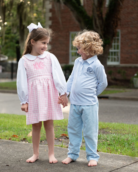 Two young children standing barefoot on a sidewalk outdoors, holding hands. The girl wears a pink and white gingham dress with long white sleeves and a white bow in her hair, and the boy wears a light blue gingham shirt and light blue pants.