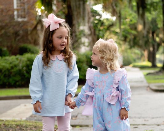Two young girls holding hands on a tree-lined path, wearing matching light blue embroidered dresses with pink accents. The girl on the left wears a large pink bow in her hair, and both dresses feature pink floral embroidery and ruffle details.
