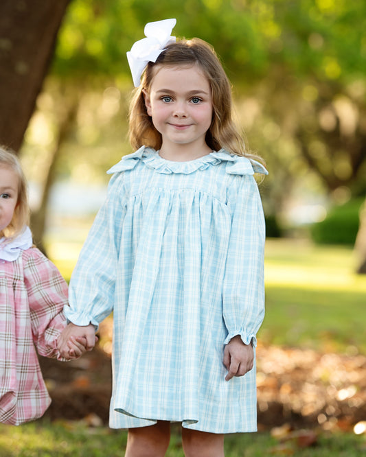 Two young girls holding hands, one in a light blue checkered dress with a white bow, the other in a pink dress, outdoors.