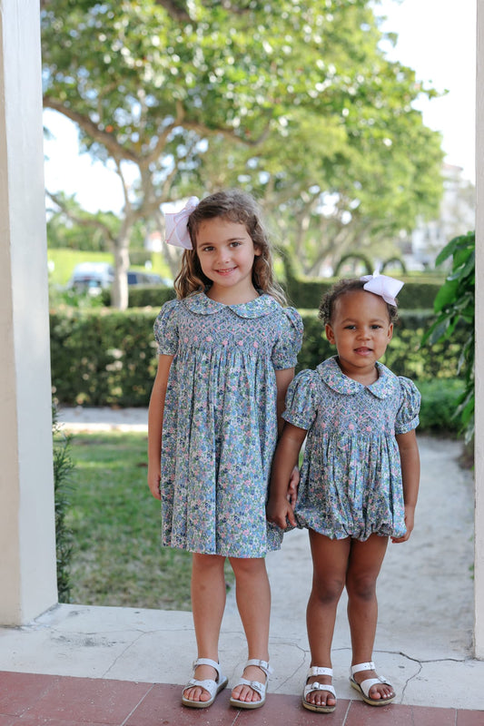 Two young girls in a matching floral dress and bubble, standing outdoors with greenery in the background.