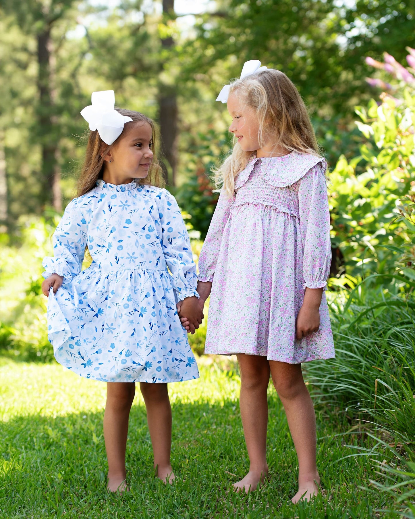 Two young girls in floral dresses standing in a grassy area with trees in the background