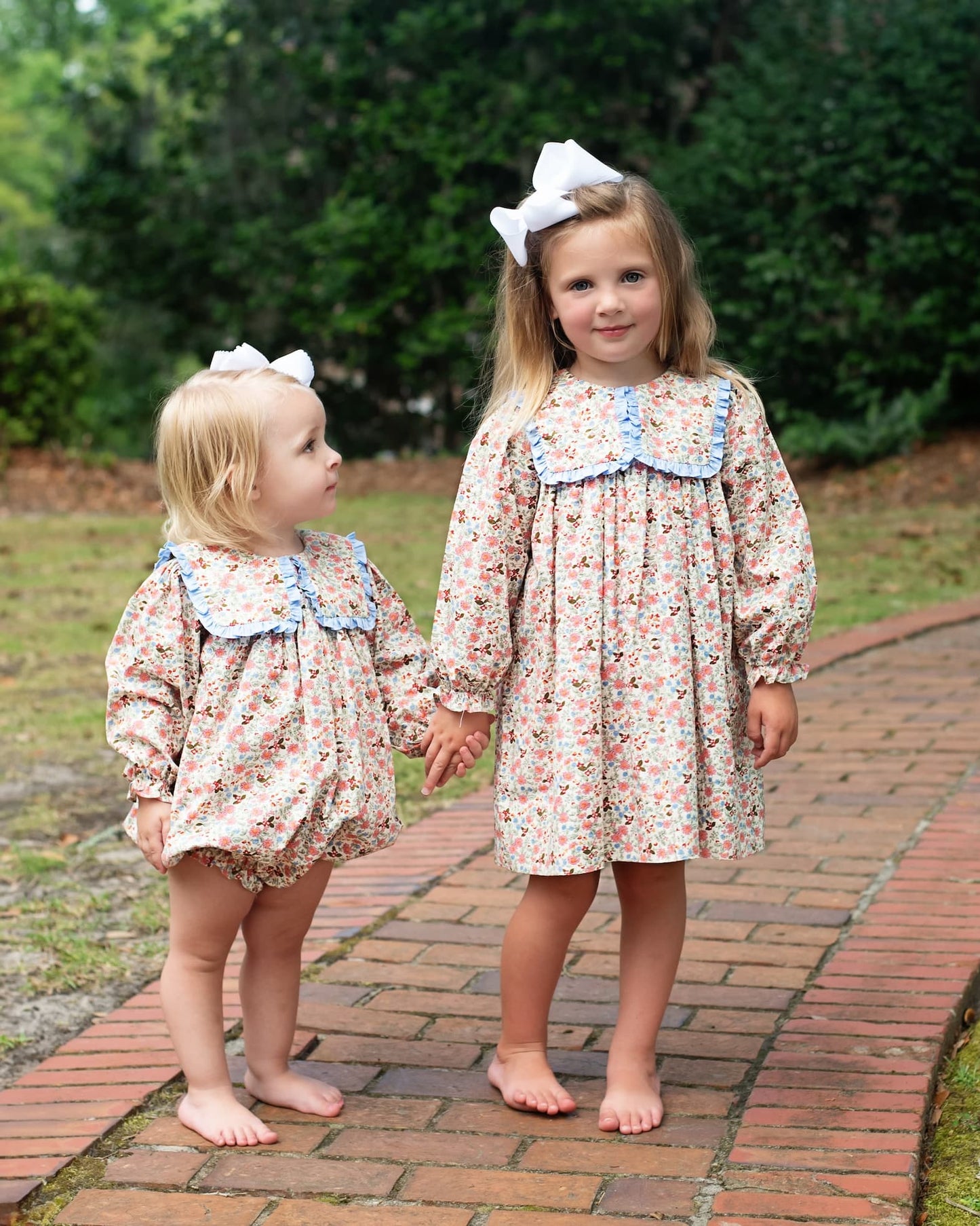 Two young girls in floral dresses standing on a brick path with greenery in the background.