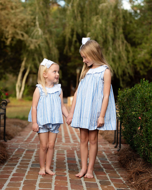 Two young girls in matching blue and white outfits standing on a brick path with greenery in the background.