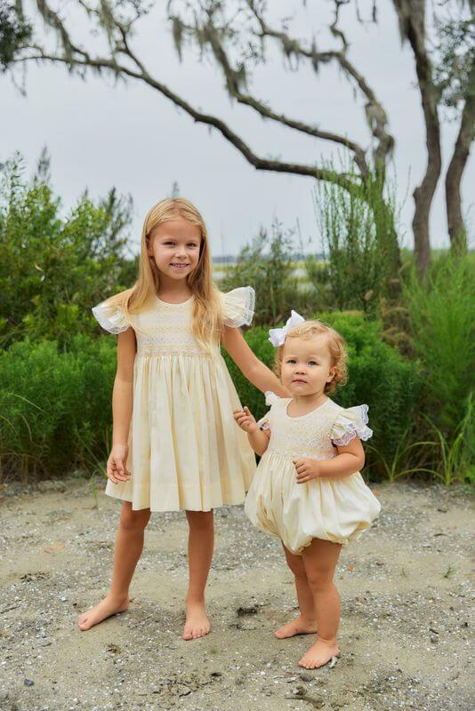 Two young girls in matching cream-colored dresses standing outdoors with greenery in the background.