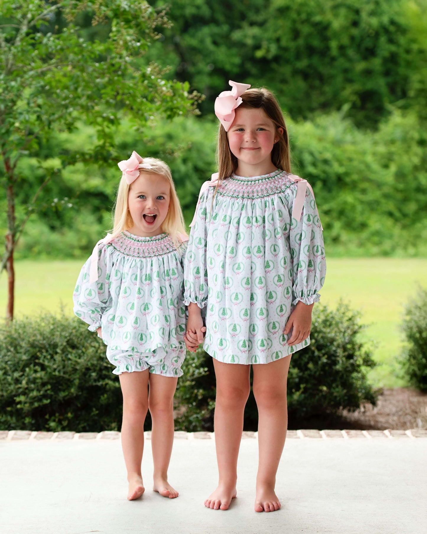 Two young girls in matching dresses standing outdoors with greenery in the background
