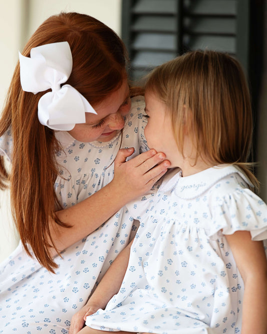 Two young girls in matching dresses with white bows sitting close together.