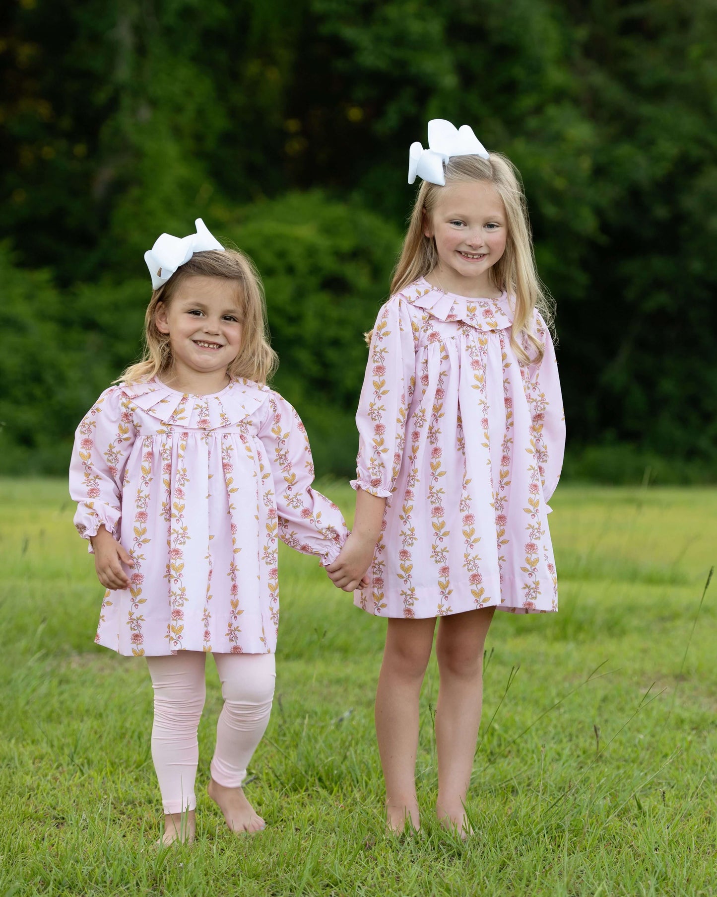 Two young girls in matching floral dresses standing in a grassy field.