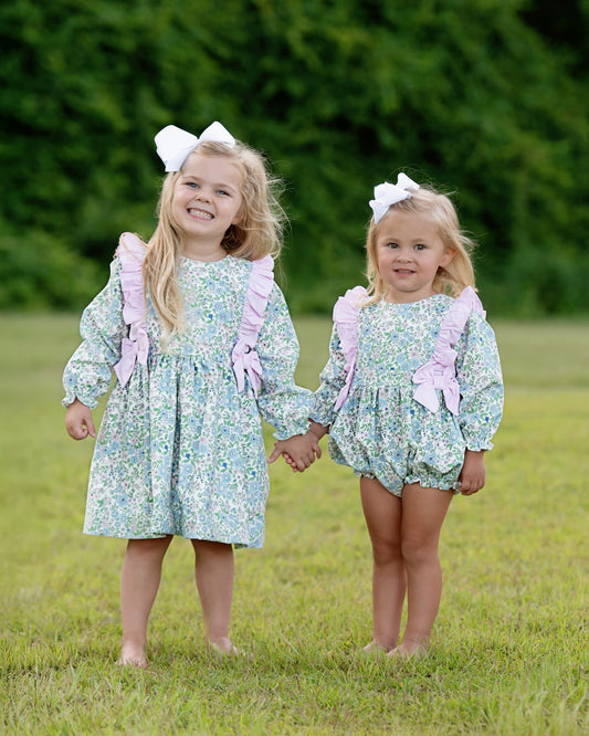 Two young girls in matching floral outfits with pink bows standing in a grassy field.
