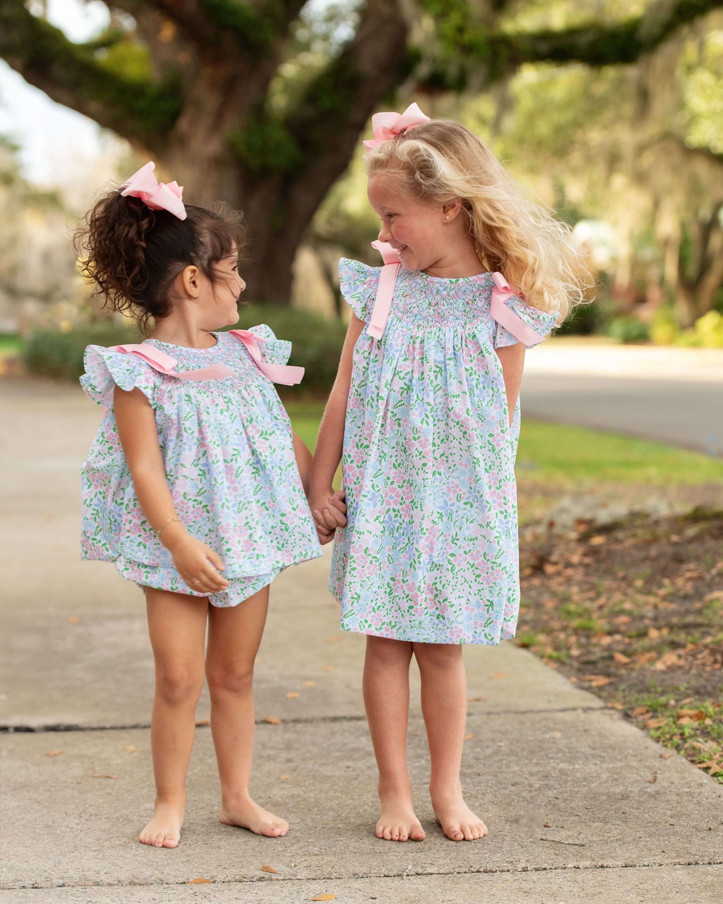 Two young girls in matching floral outfits with pink bows standing on a path outdoors.