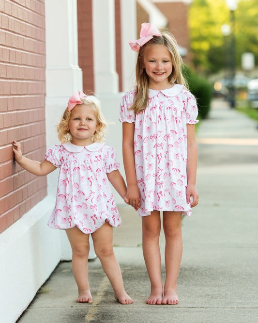 Two young girls in matching pink dresseand bubble standing on a sidewalk.