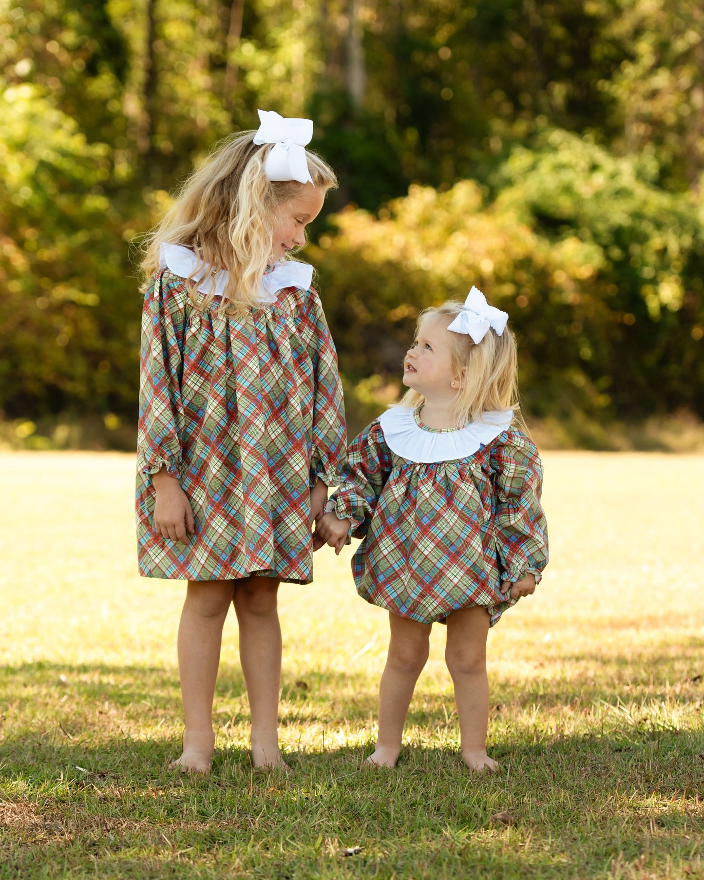Two young girls in matching plaid dresses with white bows standing outdoors.