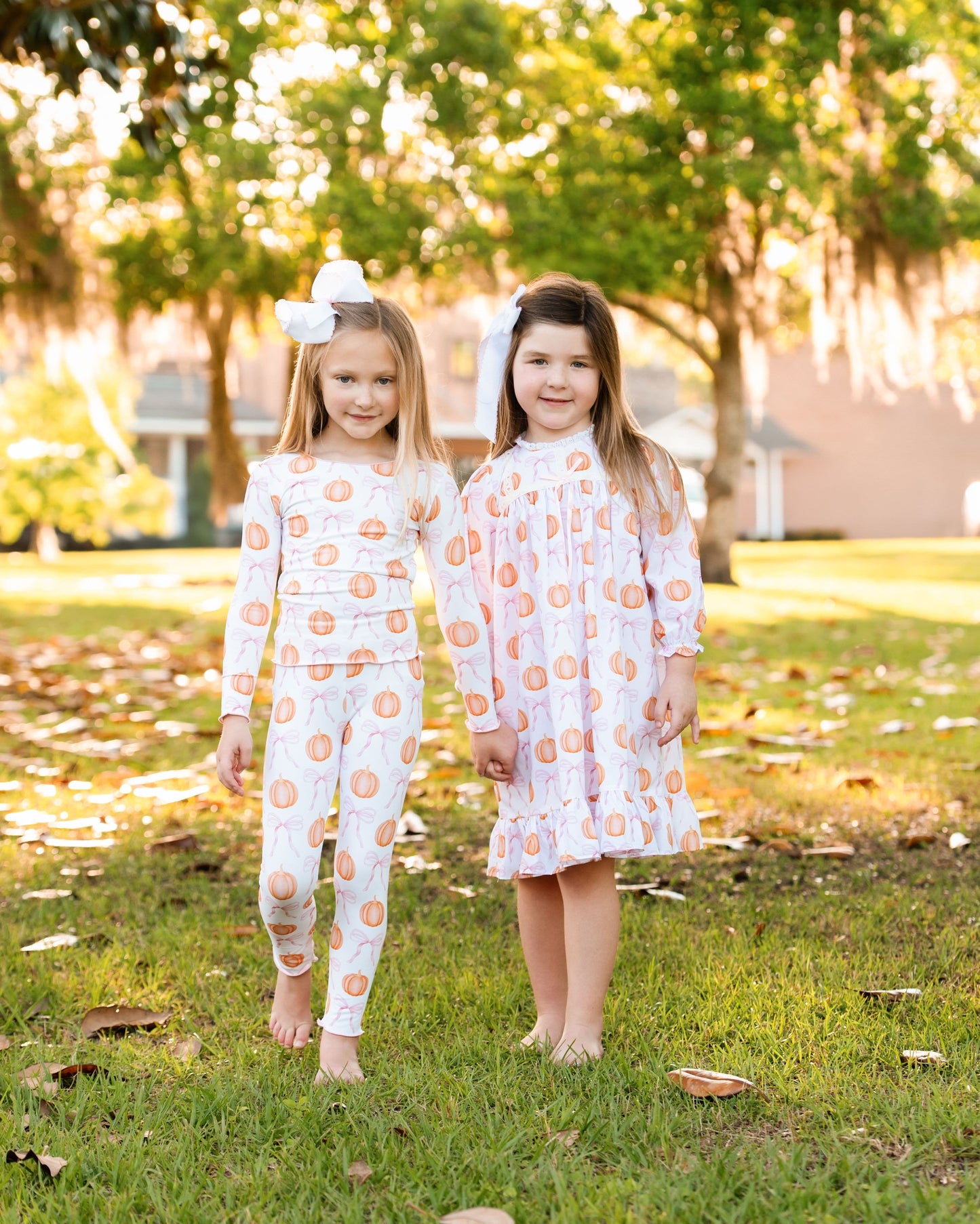 Two young girls in matching pumpkin nightgown and pajama standing on grass with trees in the background.